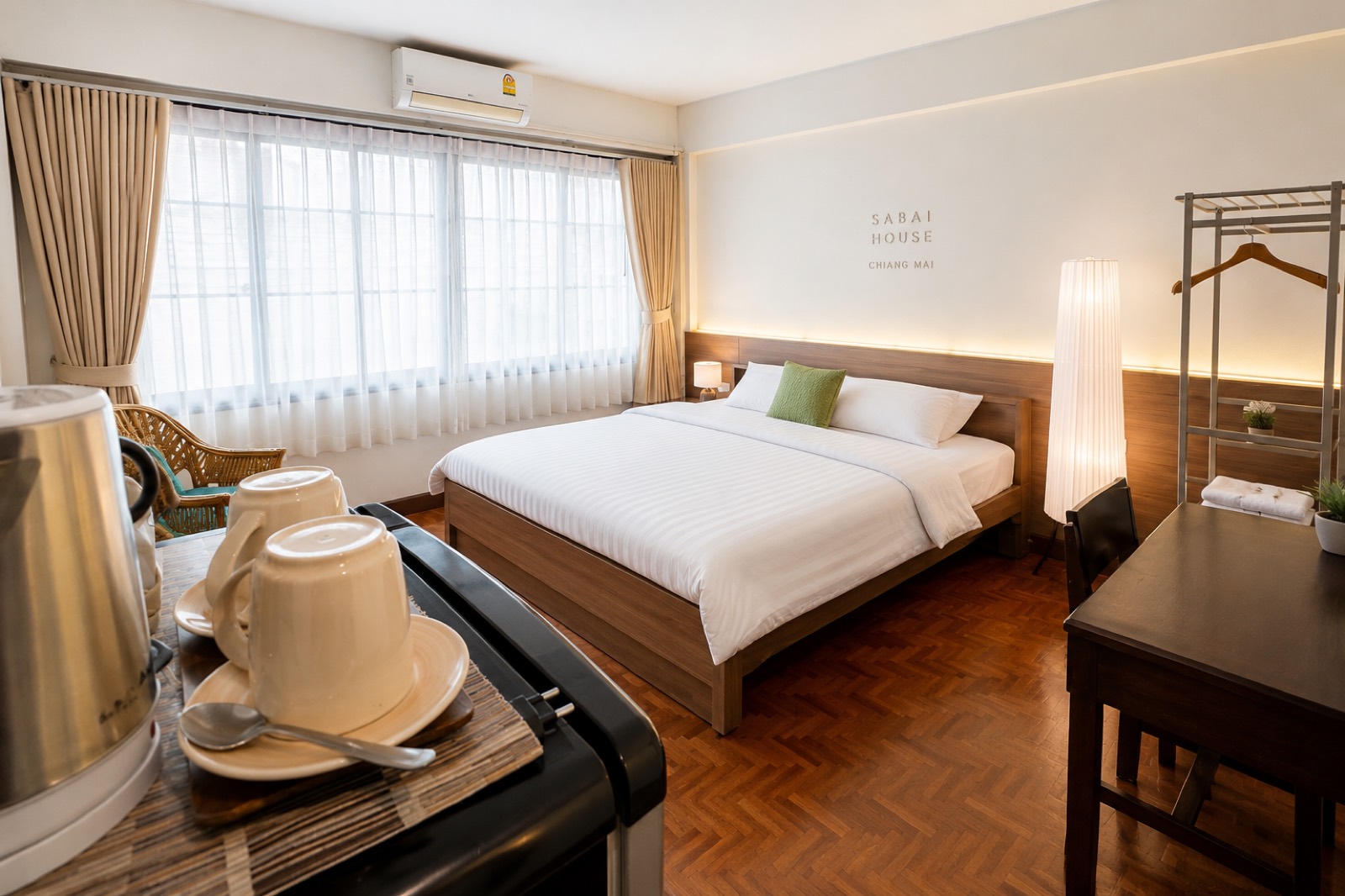 A Sabai House guest room &mdash; a wide bed with a wood headboard and warm reading lamps, a desk by the window, and Sabai House &middot; Chiang Mai lettering on the wall.