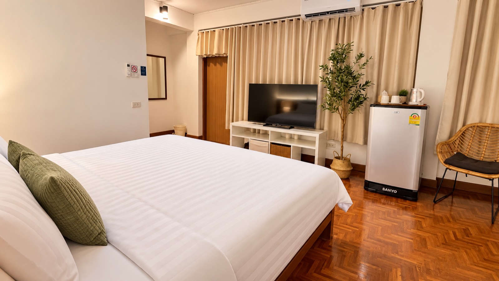 A Sabai House room &mdash; white bedding with sage cushions, a potted olive tree, herringbone parquet floor and a cane chair by the window.