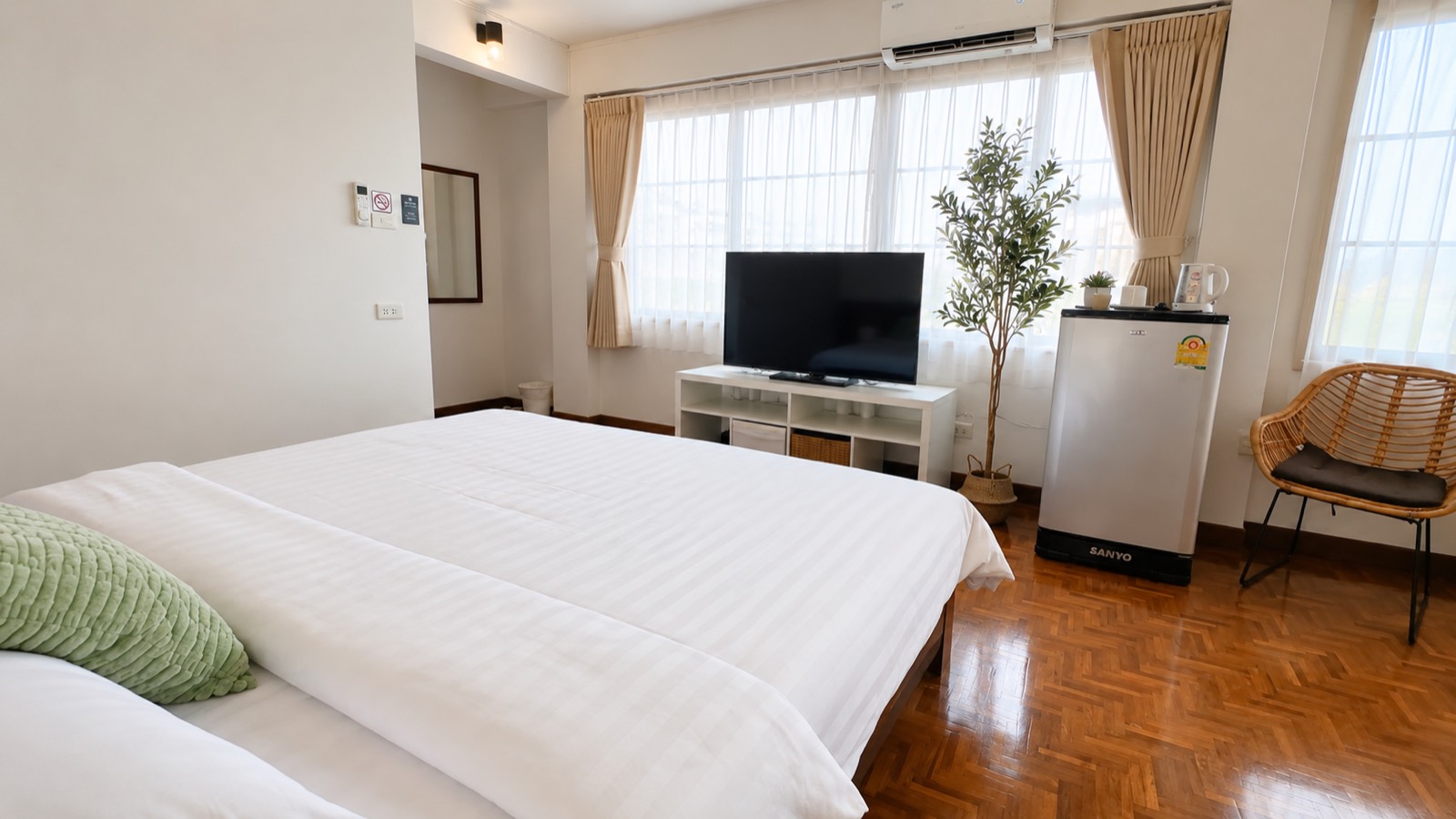 A bright corner room at Sabai House &mdash; twin beds together, windows on two sides, a cane chair and a small fridge.