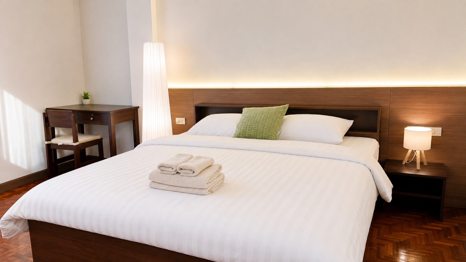 A Sabai House bed with a lit wood headboard and folded towels, a writing desk and chair to one side, warm in the evening light.